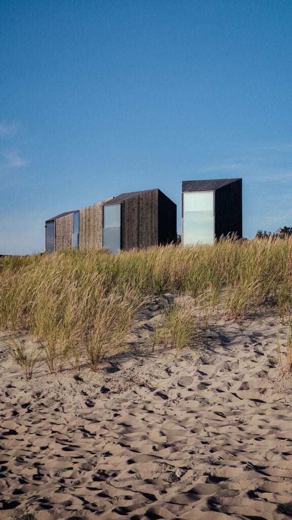 Three dark timber cabins in dune grass under a blue sky