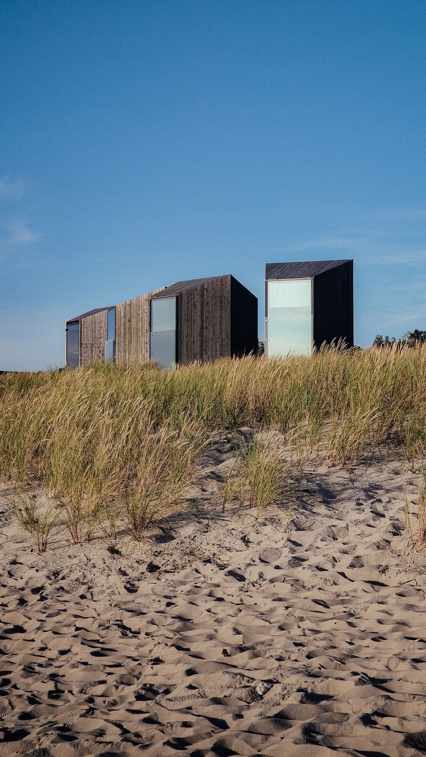 Three dark timber cabins in dune grass under a blue sky
