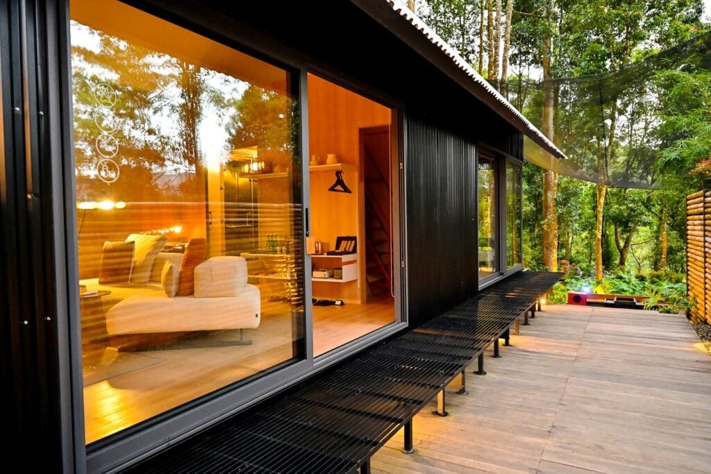 A black cabin with a glass wall and a warm lit interior beside a timber deck in the forest