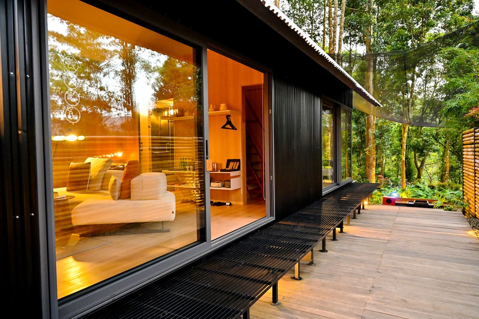 A black cabin with a glass wall and a warm lit interior beside a timber deck in the forest