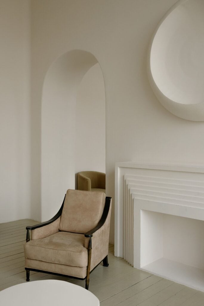A cream arched interior with a lounge chair beside a stepped fireplace