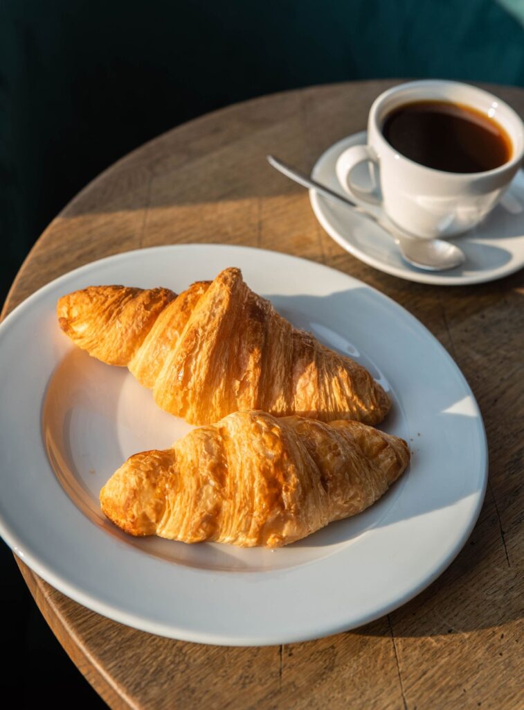 Two butter croissants and a black coffee on a wooden café table in morning sun