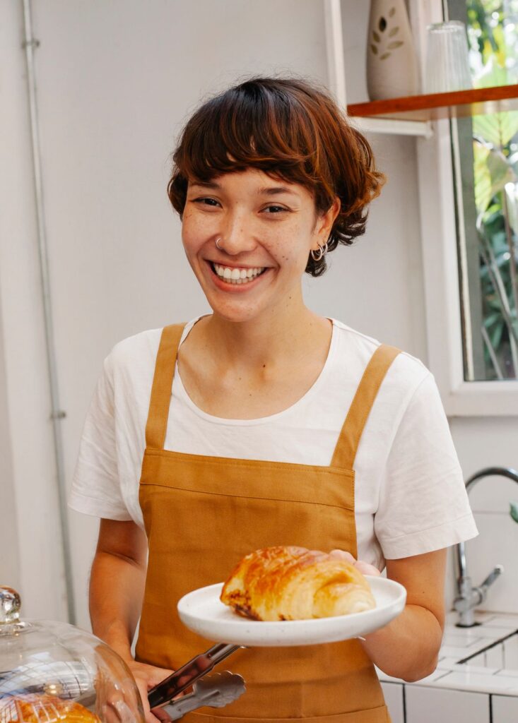 Noor, front of house, smiling in a mustard apron holding a croissant