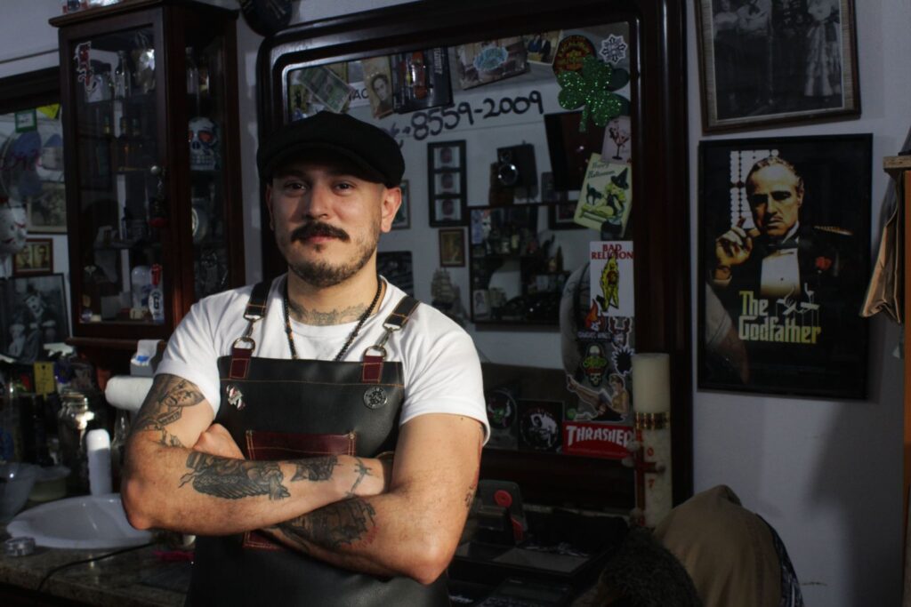 Tattooed barber in a leather apron with arms crossed inside the shop