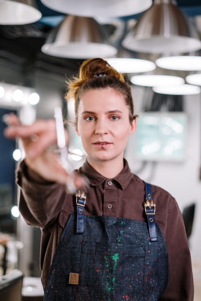 Barber in a denim apron holding scissors toward the camera