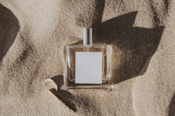 A clear glass perfume bottle with a blank label resting in sand