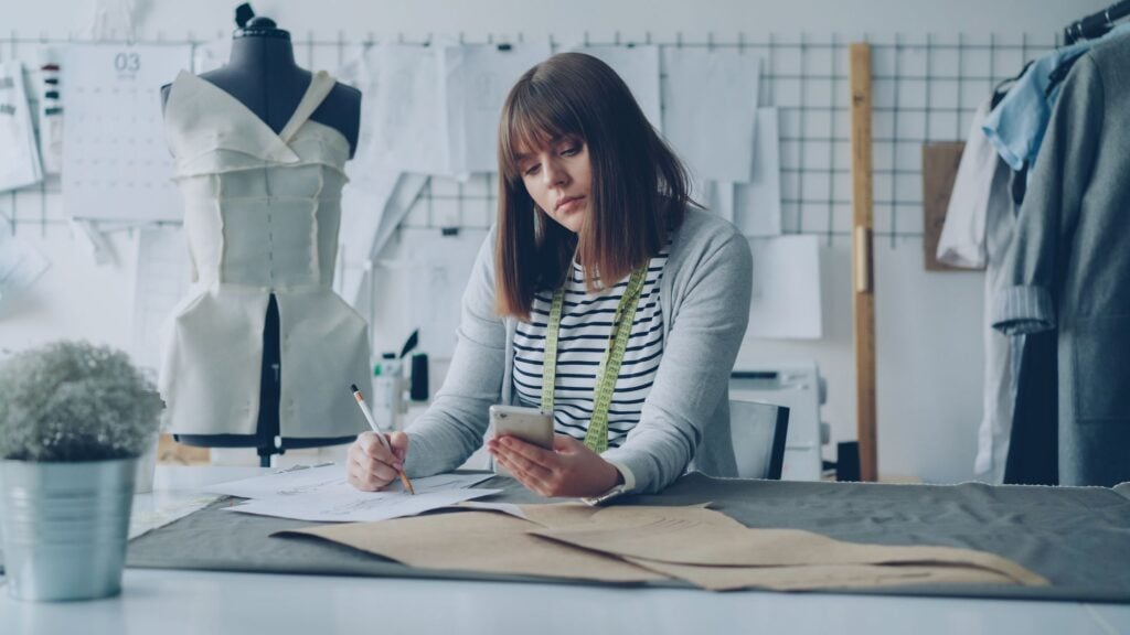 A pattern cutter drafting a pattern at a long cutting table, dress forms behind her