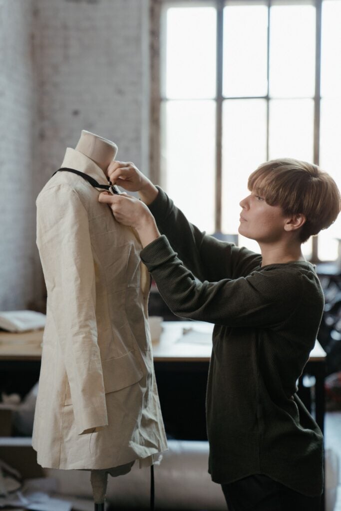 A pattern cutter fitting a natural linen jacket toile on a dress form by a tall window