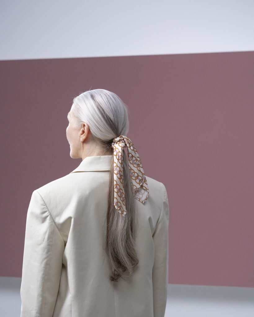 A woman in a linen suit seen from behind, a silk scarf tied in her silver hair, against a mauve wall