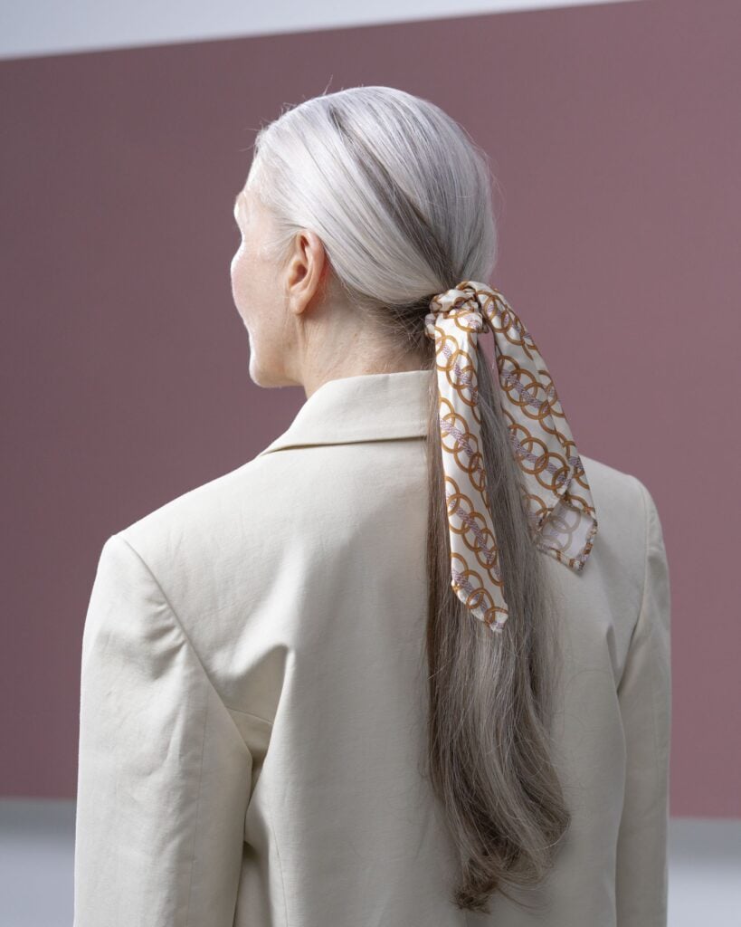 A silk scarf with a gold link print tied in long silver hair, worn with a linen blazer
