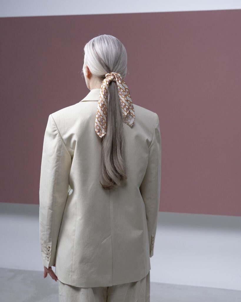 A woman with long silver hair tied with a silk scarf, wearing a natural linen suit against a mauve wall